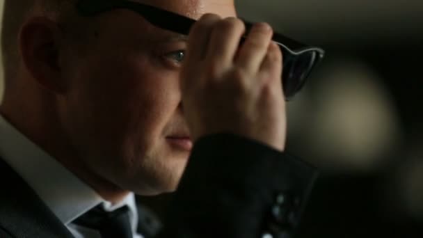Elegant businessman putting on sunglasses on the dark background - 映像、動画