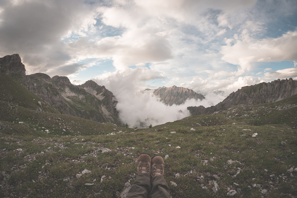 Menselijke voeten met wandelschoenen liggen op gras aan de bovenkant van de alpiene vallei met schilderachtige wolken gloeien bij zonsondergang. Ontspan terwijl kijken naar weergave. De Alpen, zomer avontuur en reizen bestemming. Getinte imag - Foto, afbeelding