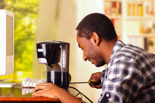 Man sitting by desk repairing small coffee maker using screwdiver, smiling happily while working - Φωτογραφία, εικόνα
