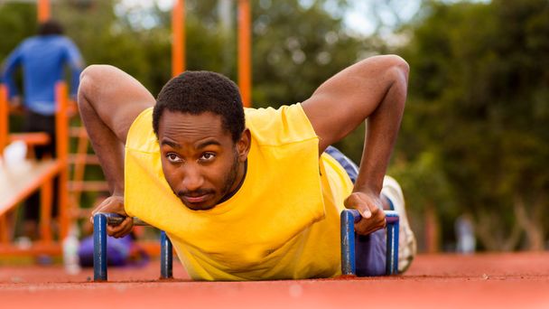 Man wearing yellow shirt doing push-ups at outdoors training facility, orange athletic surface and green trees background - Фото, зображення