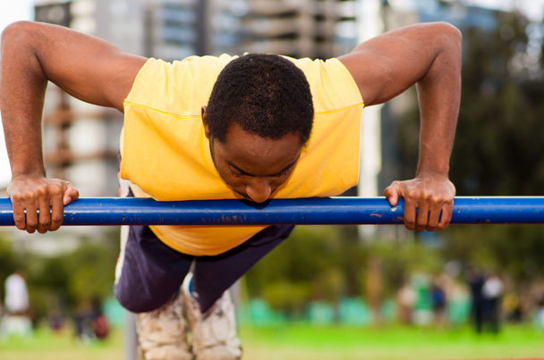 Man wearing yellow shirt and blue shorts doing static strength excercises hanging from pole, outdoors training facility with orange athletic surface, blurry people background - Zdjęcie, obraz