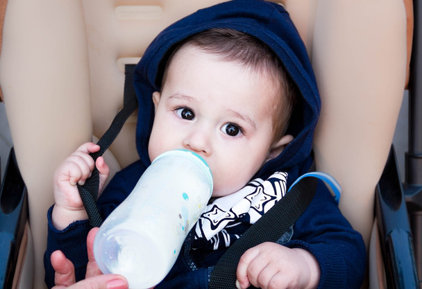 bebê pequeno feliz sentado em um carrinho de bebê e bebendo de garrafa de leite, em roupas de inverno, o peito de comida conceito
 . - Foto, Imagem
