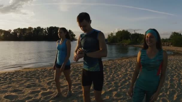 A group of athletes - two girls and a guy runs on beach, near river at sunset, use gadget for measuring heart rate - Footage, Video