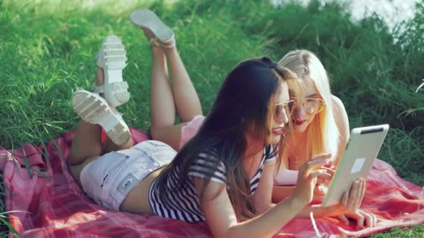 Brunette and blonde lying on blankets on the grass and using the tablet. 4k - Materiał filmowy, wideo