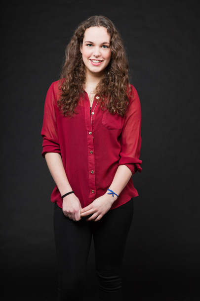 Smiling pretty girl with long brown curly hair. Fashion studio portrait isolated against black background. Wearing red shirt. - Fotó, kép