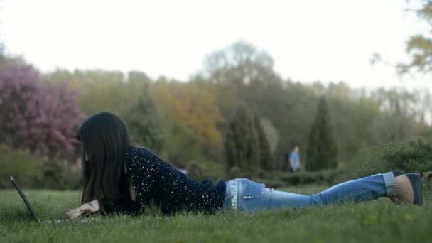Portréja egy fiatal nő, aki a zöld fűben fekve és gépelés-ra pc - Felvétel, videó