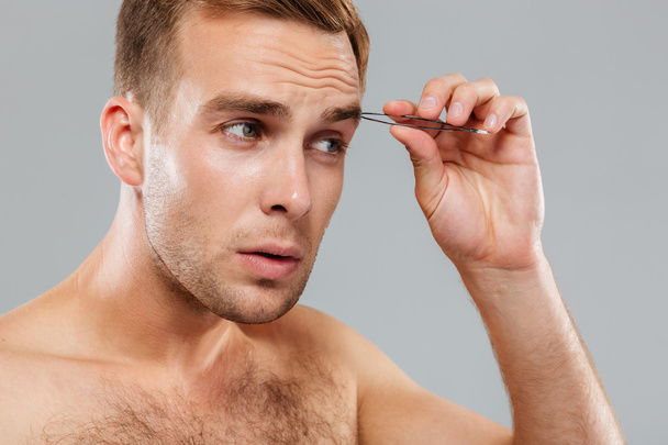 Hansome serious young man removing eyebrow hairs with tweezers - Φωτογραφία, εικόνα