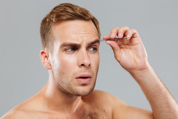 Closeup of concentrated young man removing eyebrow hairs with tweezers - Foto, Bild