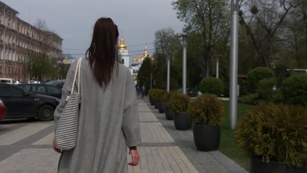 Widok z tyłu stylowe kobiece turysty, w szarym płaszczu i z torby paski podziwia piękne architektonicznych budynku podczas chodzenia w obcym mieście - Materiał filmowy, wideo