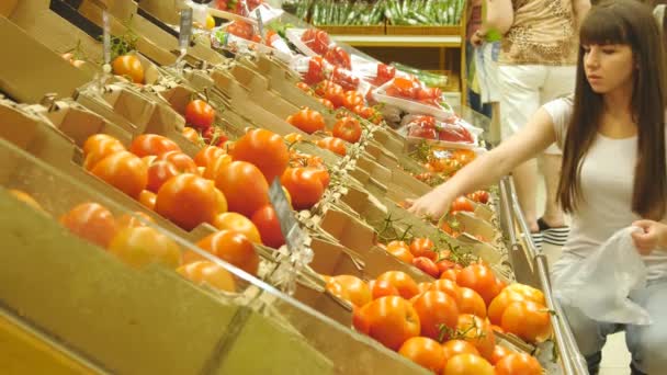 Joven chica bonita está eligiendo tomates en un supermercado y hablando. Atractiva mujer seleccionando tomates rojos maduros frescos en el departamento de productos de comestibles
. - Metraje, vídeo