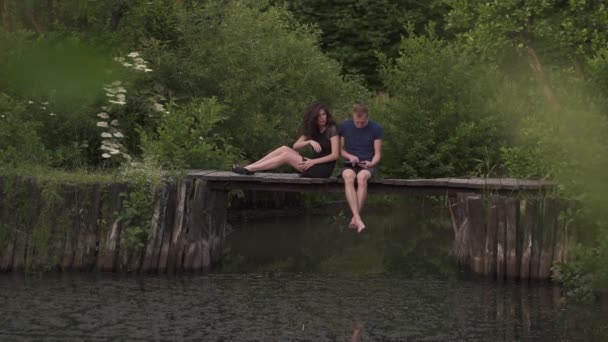 Ζευγάρι που κάθεται σε ένα ξύλινο ποντονίου σε ένα ποτάμι το καλοκαίρι. Χρησιμοποιώντας μια ψηφιακή δισκίο και σύζυγος άντρα είναι θυμωμένος και αναστατωμένος σχετικά με λιγότερη προσοχή - Πλάνα, βίντεο