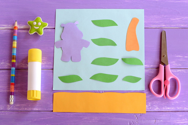 Hippo, leaves, palm tree trunk cut from colored paper. Set for children craft projects. Paper card hippo craft. Glue stick, scissors, pencil, eraser on wooden background. Top view  - Photo, Image