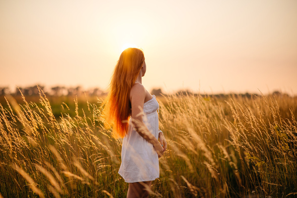 Young beautiful red-haired girl in a field at sunset - Foto, imagen