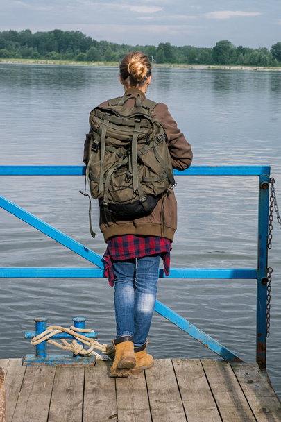 rear view of blonde girl with backpack on handrail watching river - Foto, afbeelding