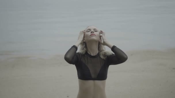 Front view of sexy blonde woman wearing a black elegant swimsuit posing on the beach over sea background - video in slow motion. - Imágenes, Vídeo