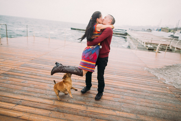 Ein junges glückliches Paar küsst sich und spielt mit dem Hund auf dem verregneten Liegeplatz im Herbst. Hintergrund Meer - Foto, Bild