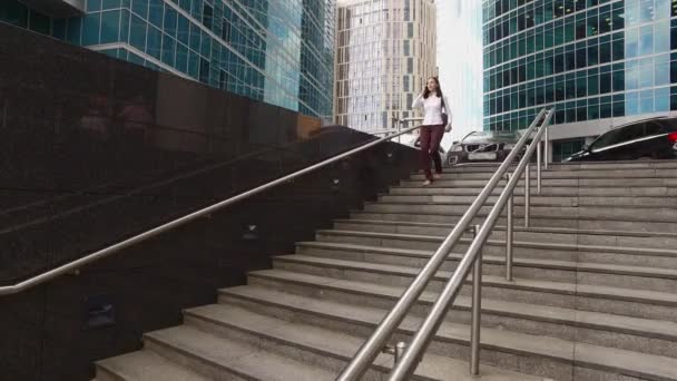 Beautiful girl is going down the modern stairs and talking on the phone near luxury offices and skyscrapers (steadicam shot) - Materiaali, video