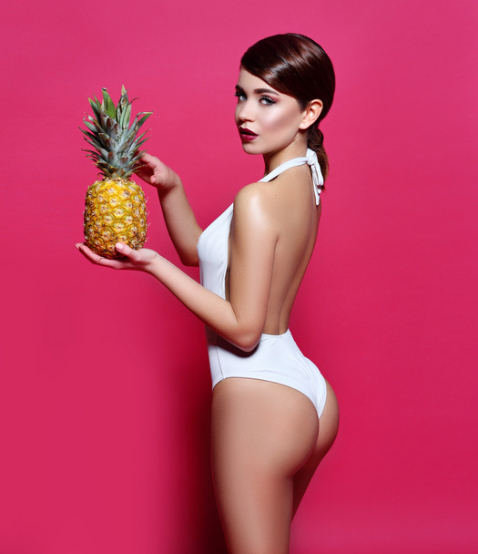 beautiful sexy young girl standing on a pink background with  magazine cover with perfect body with bronze tanned skin holding a pineapple cocktail smiles a lovely smile perfect photos for advertising - Foto, afbeelding