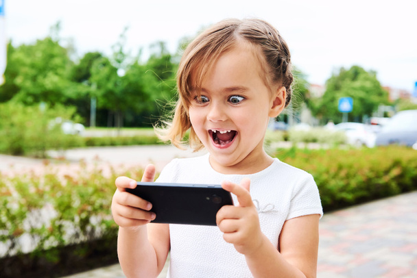 Adorable expressive little girl with a smartphone outdoors - Φωτογραφία, εικόνα