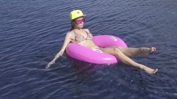 Mooie jonge vrouw die het dragen van bikini, gele hoed en paarse zonnebril zittend op een roze opblaasbare ring in zwembad op een zonnige dag. - Video