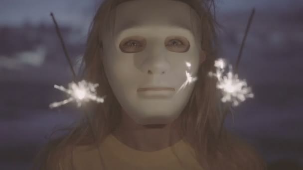 Closeup a sebhelyes szőke nő visel fehér maszkot csillagszóró tűzijátékok a tengerparton Alkonyat. - Felvétel, videó