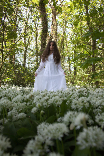 Hermosa mujer en vestido blanco largo de pie en un bosque sobre una alfombra de flores blancas
 - Foto, Imagen