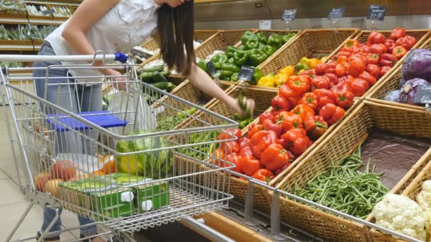 Eine junge Frau schiebt einen Einkaufswagen durch die Gänge des Supermarktes. Mädchen wählen frisches Gemüse im Supermarkt und legen es in den Einkaufswagen. Frau kauft Paprika im Geschäft - Filmmaterial, Video