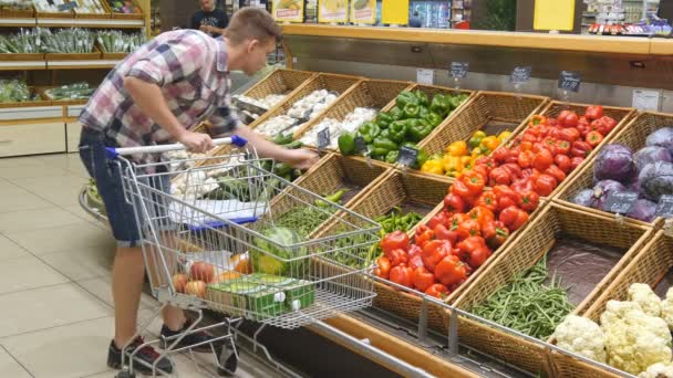 Mladý muž tlačí vozík podél uliček s potravinami v supermarketu. Chlap, výběr čerstvé zeleniny v obchodu s potravinami a jejich uvedení v nákupním vozíku. Chlapec, nákup papriky v obchodě - Záběry, video