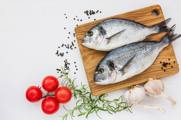 Świeże ryby dorado na deska do krojenia z czosnkiem, rozmarynem i pomidory na biały stół. Widok z góry - Zdjęcie, obraz