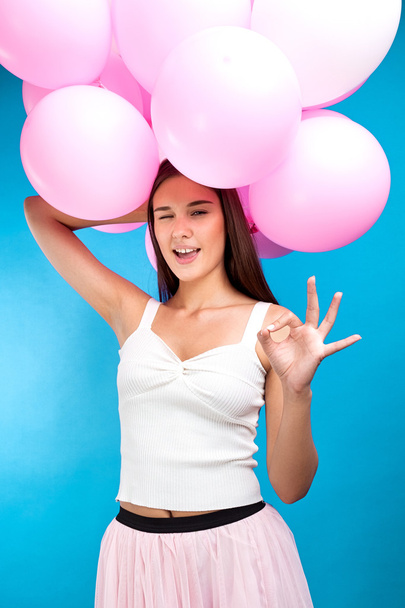 Portrait of pretty brunette holding pink balloons like crown while winking at camera with ok gesture - Фото, изображение