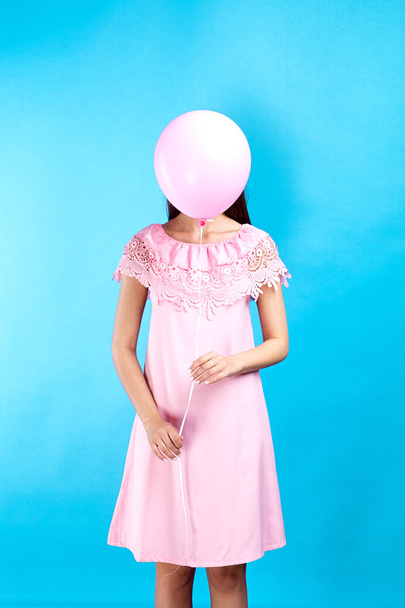 Conceptual shot of unrecognizable brunette in pink dress with pink balloon hiding her face on blue background.Isolate - Photo, Image