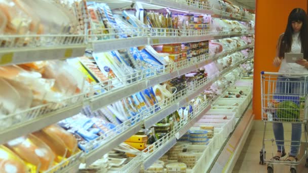 ショッピング カートの購入を持つ若い女性は冷蔵食料品スーパー マーケットでタブレット pc を使用して、買い物リストを確認してください。それから店で取って製品冷蔵庫に来る女の子 - 映像、動画