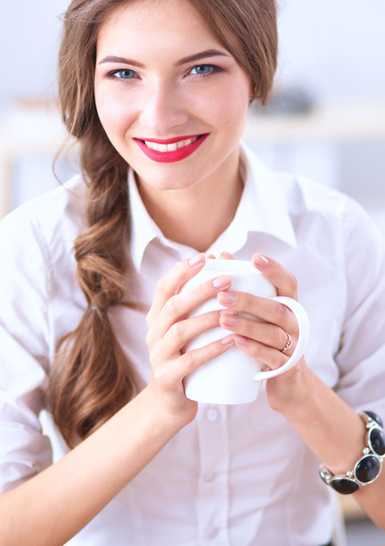 Young businesswoman sitting on the desk with cup in office - Foto, afbeelding