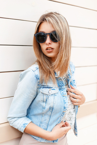 Сексуальная красивая стильная девушка в солнцезащитных очках и синей джинсовой куртке, стоящая у деревянной стены
 - Фото, изображение