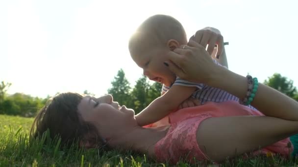 Νεαρή ευτυχισμένη μητέρα με ένα παιδί που βρίσκεται και ακουμπά σε ένα πράσινο γκαζόν σε ένα πάρκο - Πλάνα, βίντεο