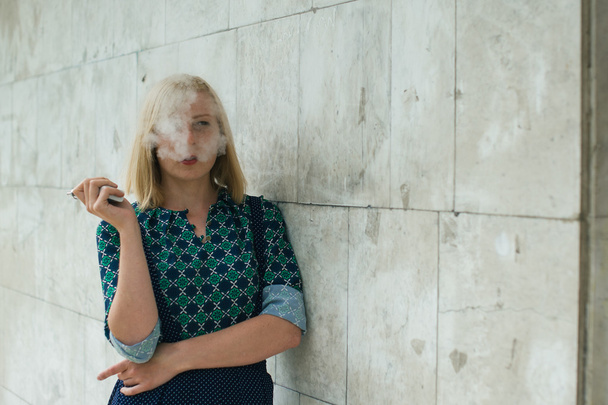 young blonde girl smokes electronic cigarette on the background wall - Fotó, kép