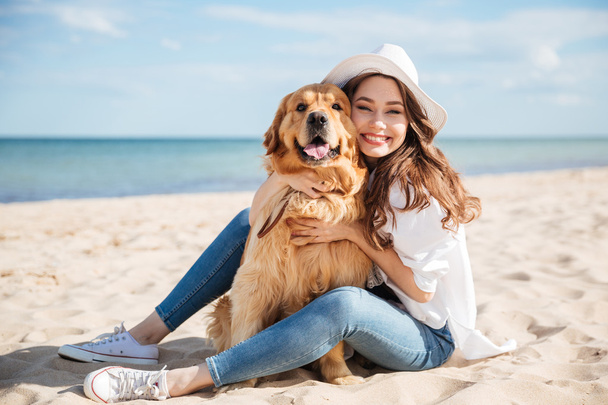 Cheerful young woman sitting and hugging her dog on beach - Photo, image