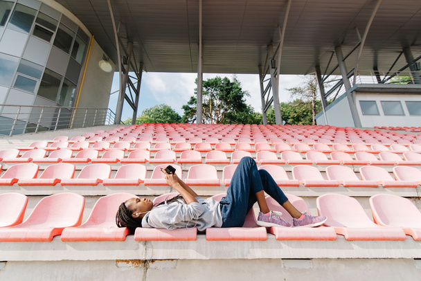 Cute african american young woman listening to music with earphones lying on a tribune at outdoors stadium - Фото, зображення