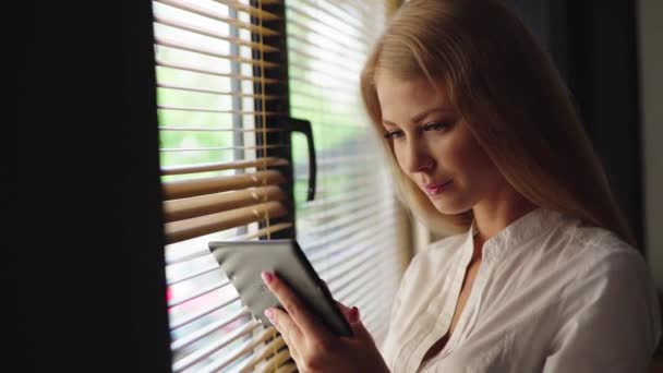 Крупним планом чарівна блондинка тримає сенсорну панель, стоячи біля вікна в сучасному інтер'єрі
 - Кадри, відео