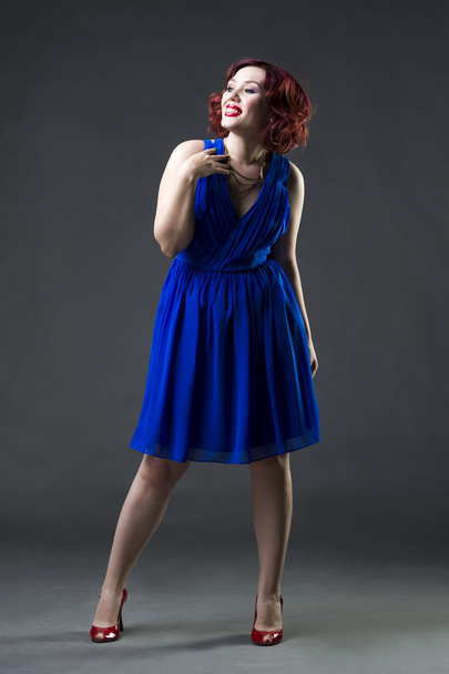 Young beautiful red-haired caucasian woman in blue dress posing in studio on gray background, professional makeup and hairstyle - Foto, Imagem