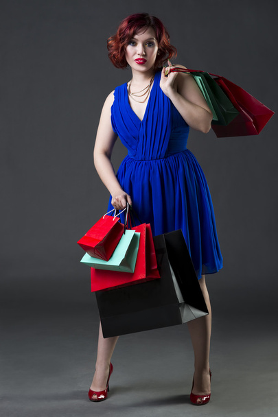 Shopaholic έννοια, νεαρή όμορφη κοκκινομάλλα καυκάσιος γυναίκα στο μπλε φόρεμα που θέτουν σε studio σε γκρι φόντο, επαγγελματικό μακιγιάζ και χτένισμα - Φωτογραφία, εικόνα
