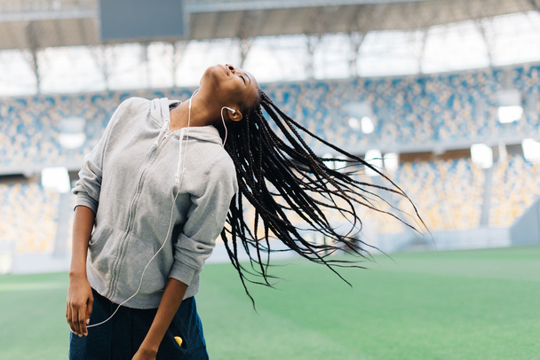 Jeune adolescente afro-américaine en sweat à capuche gris écoutant de la musique et jouant avec ses cheveux au stade
 - Photo, image