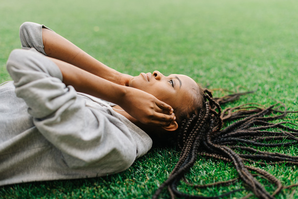 Thoughtful young african american girl lying down on the grass listening to music with headphones - Foto, Imagen