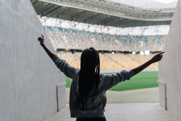 Happy african american woman standing among empty stadium celebrating, her arms raised punching the air with hands, view from back - Zdjęcie, obraz