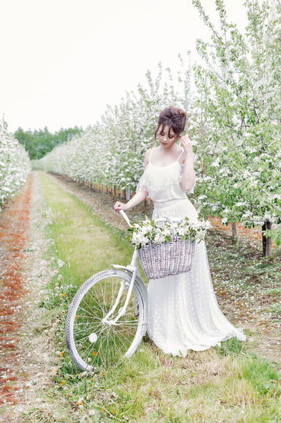 beautiful gentle lovely girl bride in a white dress standing with his bicycle in blossoming apple orchard - Fotografie, Obrázek