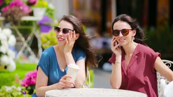 Δύο νεαρά κορίτσια χρησιμοποιούν έξυπνο τηλέφωνο στην υπαίθρια καφετέρια. Δύο γυναίκες μετά τα ψώνια με τσάντες που κάθονται σε openair cafe με καφέ και τη χρήση smartphone - Πλάνα, βίντεο