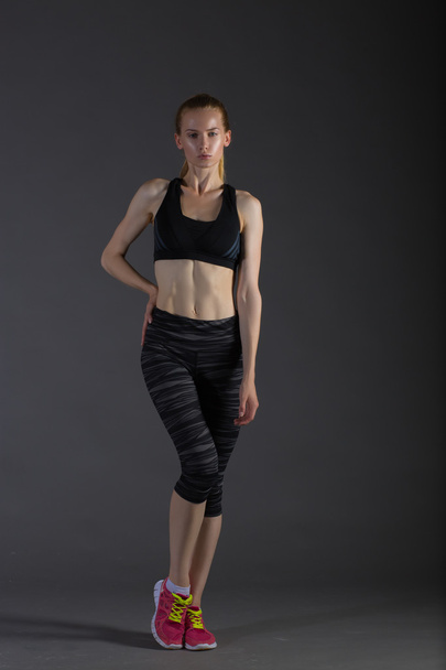 Body of slim female in activewear doing posin on gray low key, perfect blonde - Фото, изображение