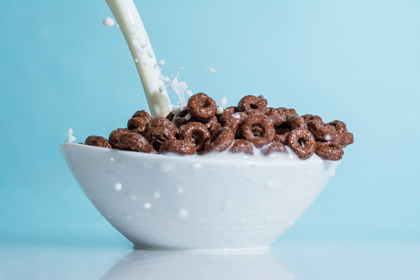 Milk stream jet pouring into a cup with chocolate flakes in the form of rings, splashes of milk on a light blue background - Fotografie, Obrázek