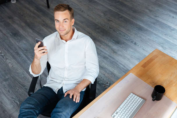 Smiling relaxed young businessman sitting and using smartphone in office - Фото, зображення