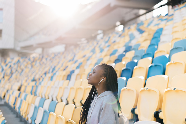 Muchas sillas de plástico azules y amarillas y una chica afroamericana solitaria con auriculares en el estadio
 - Foto, Imagen